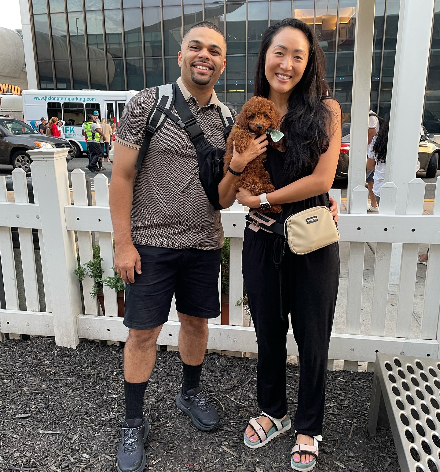 Two adults outdoors beside a white fence with a small brown puppy