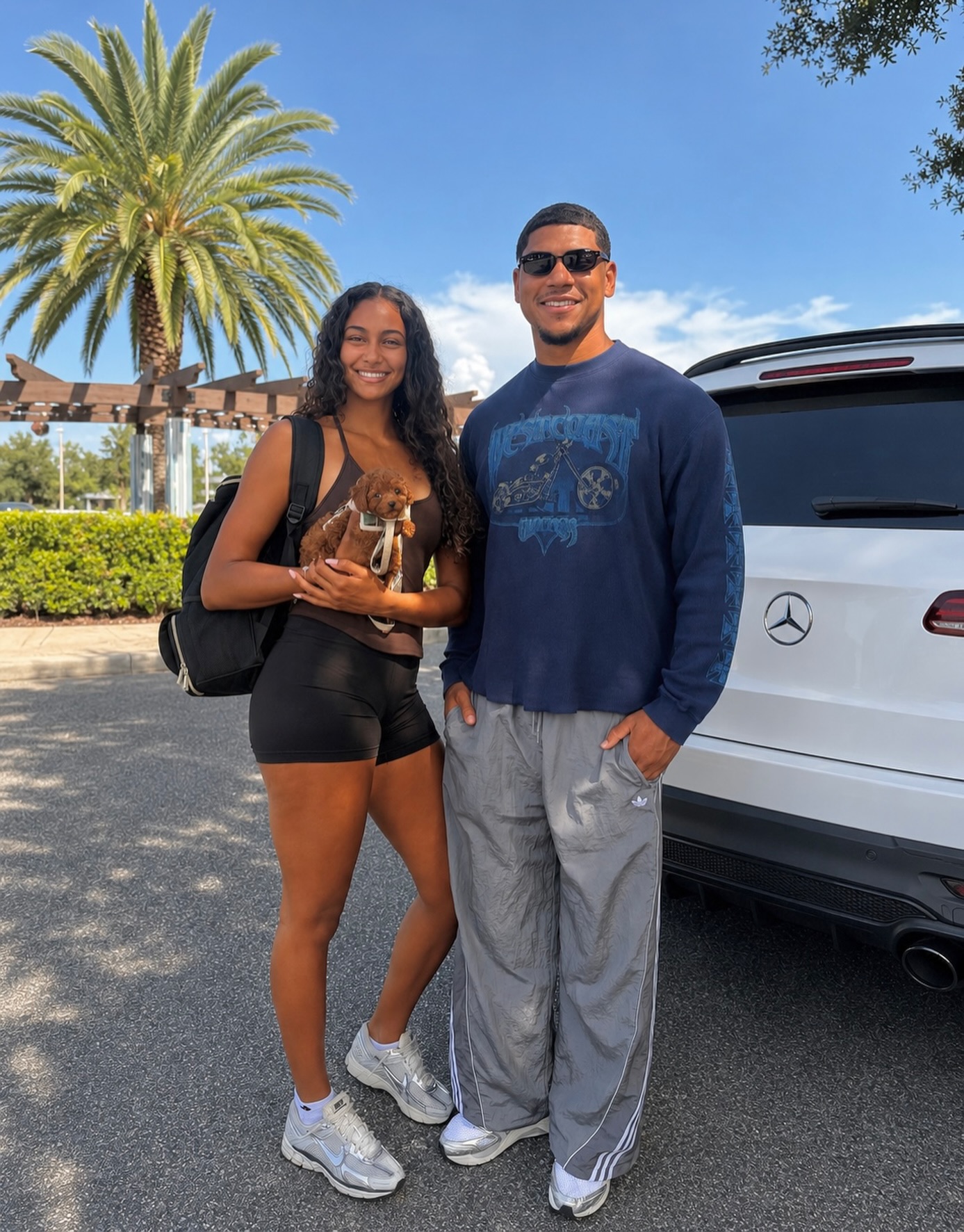 Two adults with a small brown puppy standing near a white vehicle outdoors